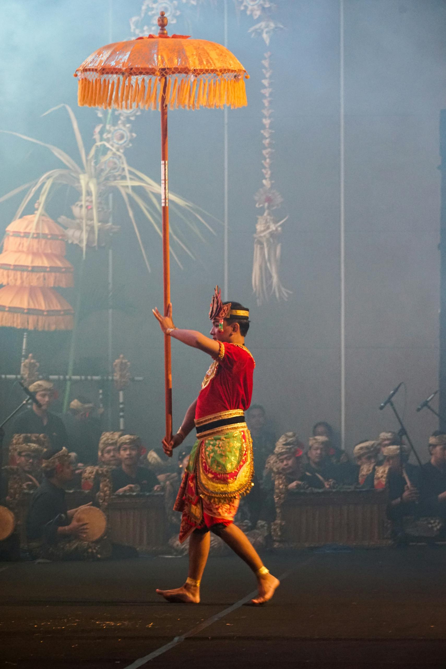 A dancer in vibrant costume performs a traditional Balinese dance with an ornate umbrella.