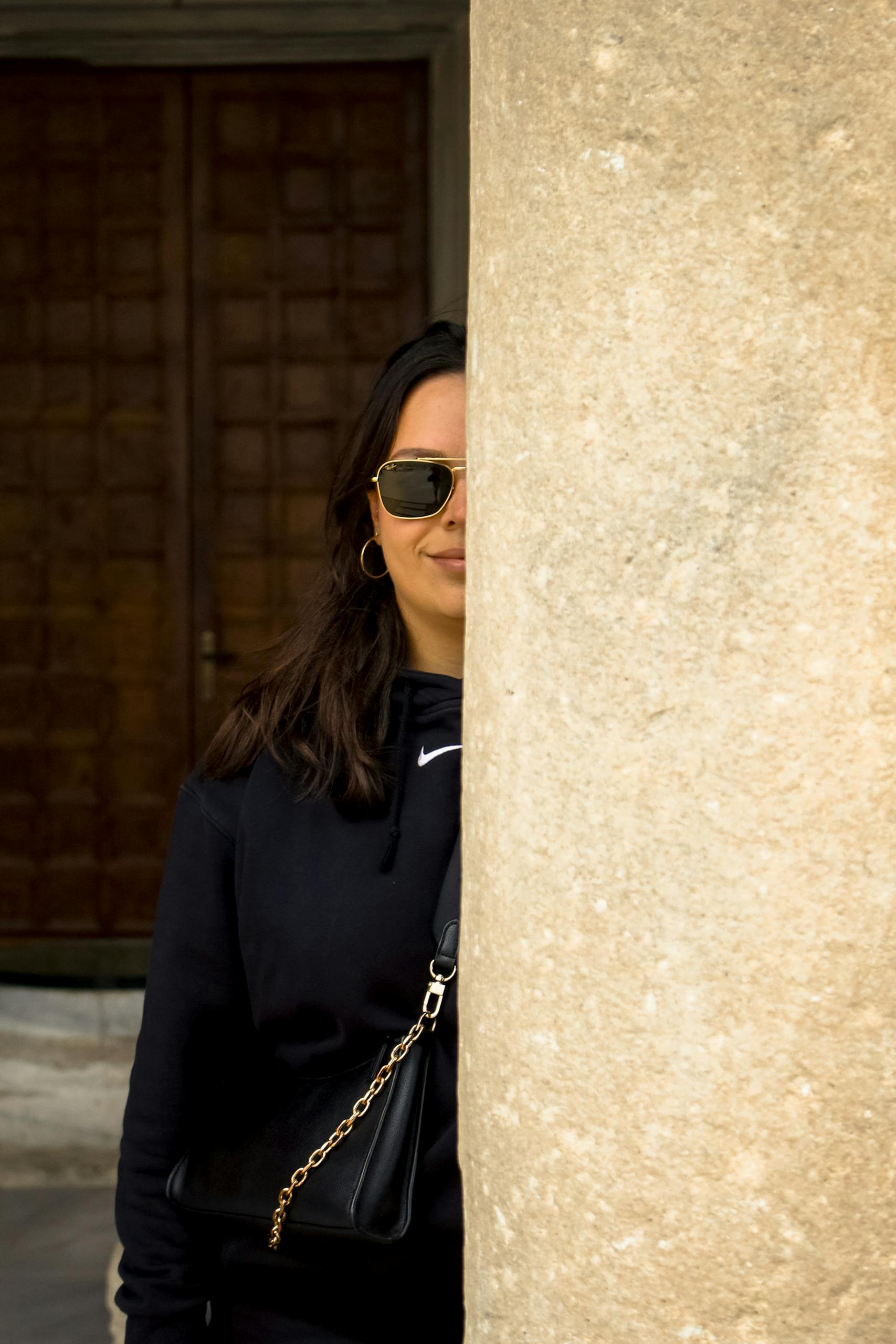 Stylish woman in sunglasses peeking from behind a column in an urban setting.